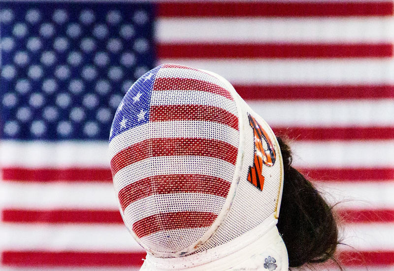 A fencer in a mask painted like the American flag stands in front of a hanging flag