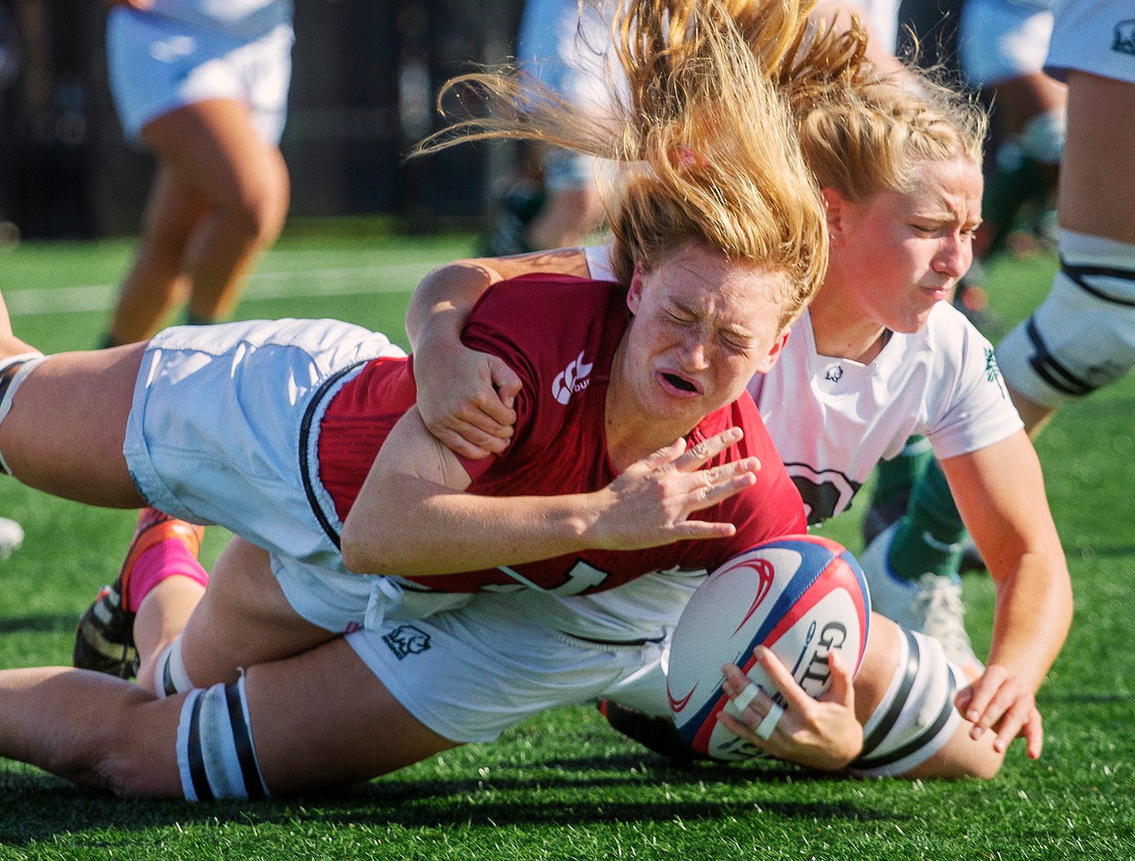 A women's rugby player in maroon dives with the ball as a defender in white drags her down